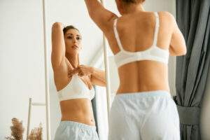 Woman in white bra examining her breast in the mirror for signs of hematoma post-surgery.