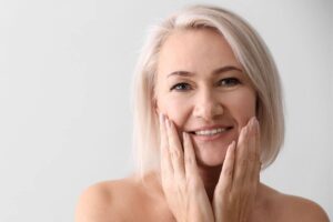 Smiling woman touching face gently, reflecting natural-looking rejuvenation after a deep plane facelift recovery.